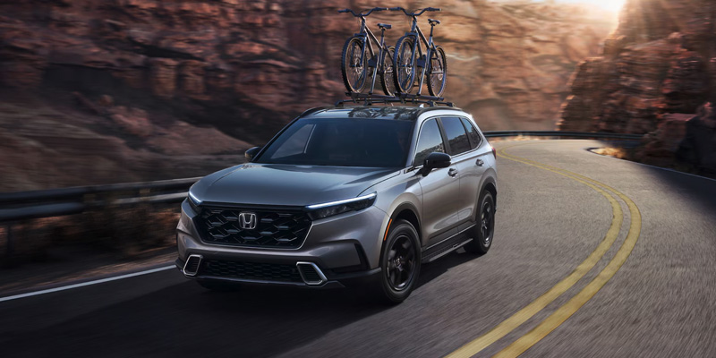 A grey 2025 Honda CR-V Hybrid with bikes on the roof driving down a road